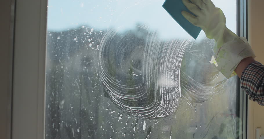 Woman in rubber gloves sprays window cleaner from a spray bottle onto the windows glass. Housewife hands in rubber gloves cleaning windows close up. Cleaning glass window with a cloth special rag blue