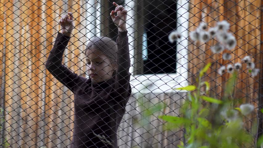 Unhappy child standing behind of fence. Close-up of a child girl standing next to wire fence holding hands on it, looking sad