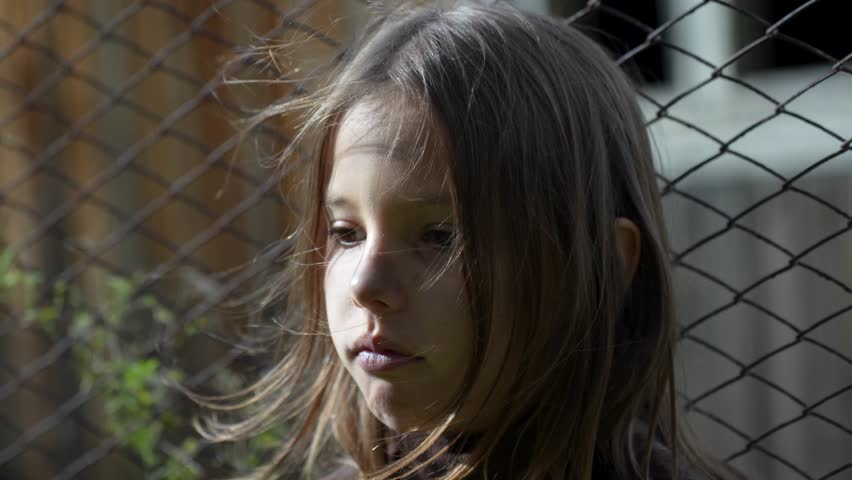 Close-up of unhappy child standing at wire fence.  Little girl behind wire fence looking down