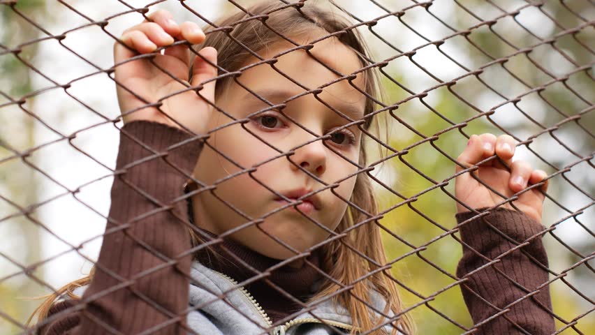 Close up of lonely girl outside the fence. Little girl looking through a wire fence