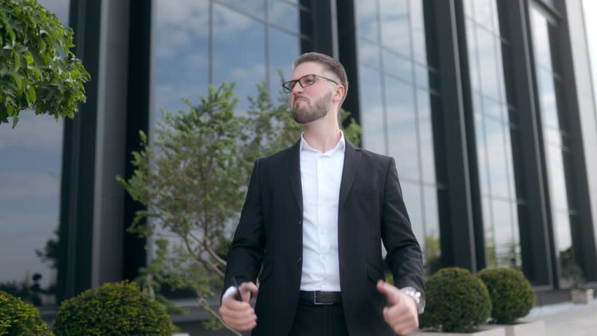 A joyful professional in glasses and a suit celebrates outside a corporate office building. The image captures the moment after he received good business news, showing his excitement.