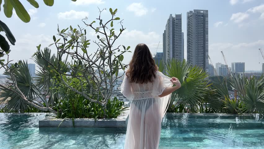 young beautiful girl throws off white transparent pareo goes to the pool beautiful figure black swimsuit pool on the roof of the house against the backdrop of skyscrapers Vietnam Ho Chi Minh City