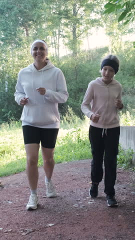 Vertical video. A cheerful mother and her daughter enjoy a refreshing morning run in a serene park, surrounded by lush greenery, embodying a healthy lifestyle concept