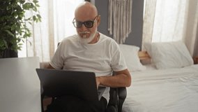 Man working on a laptop in a bedroom with a bed in the background and a plant nearby, wearing white t-shirt and glasses - Powered by Shutterstock - Get 15% off with code: PIKWIZARD15