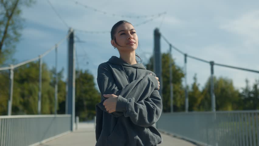 A smiling teenage girl stands in a serene park setting, her arms around herself, enjoying the peace and freedom of a carefree moment outdoors.