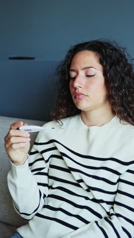 A sick woman with a fever sits on her sofa, feels sick and checks her temperature with a thermometer.   Woman feeling sick checking temperature on thermometer
