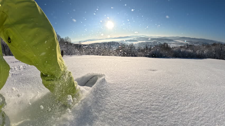 POV: Snowboarder carving through fresh snow on a sunny winter day. The snow sprays in bright sunlight as enthusiastic splitboarder rides down the snowy mountain with a joyful dog jumping by side.