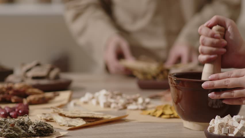 The Oriental Medicine room setting with a table filled with herbs features a herbalist pounding angelica sinensis while a herbalist in beige clothes rearranges the wrapped herbal medicine packets.