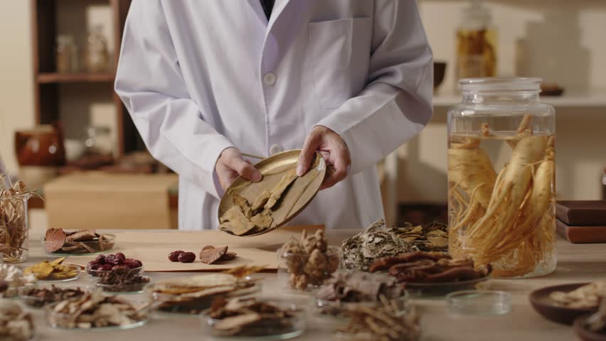 A physician places dried herb slices from a conventional scale on brown paper on a table, surrounded by other herbs. A traditional laboratory setting is appropriate for advertising natural medicines.