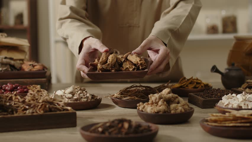 The herbalist holds up a wooden plate filled with Codonopsis. On the table are wooden plates and wooden trays filled with different herbs. Traditional clinic space suitable for herbal medicine videos.