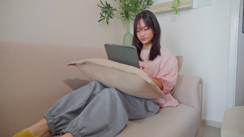 Asian young teenager woman using digital tablet in living room at home. Adorable girl sitting on sofa and smiling while holding tablet for virtual distancing studying online learning alone in house  - Powered by Shutterstock - Get 15% off with code: PIKWIZARD15