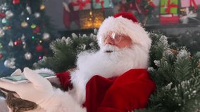 Modern Santa Claus sits in a chair relaxed using a mobile phone stroking a domestic cat among fir branches decorated with Christmas tree decorations and colorful garlands - Powered by Shutterstock - Get 15% off with code: PIKWIZARD15