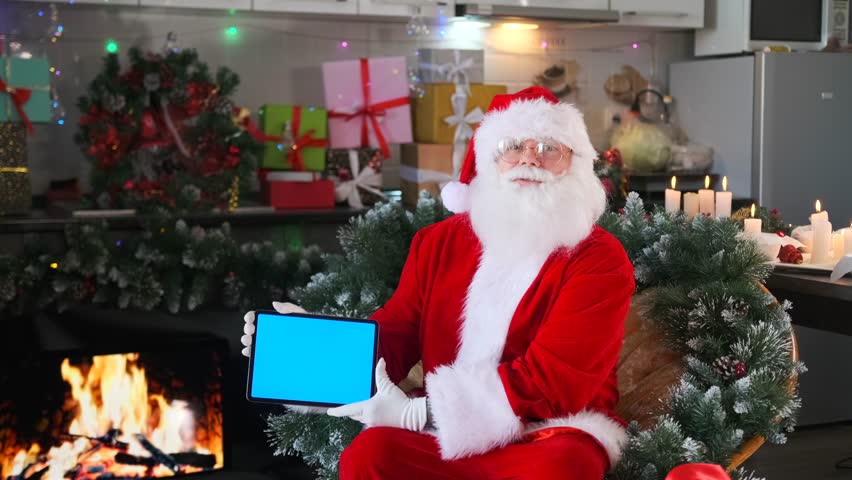 Charismatic santa holding tablet with blank screen sitting in his residence decorated with Christmas lights tree candles and gifts.