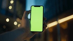 Camera view of Caucasian male's hand holding smartphone vertically. Green screen visible on display of mobile device. Cell phone with chroma key. Evening in city visible in background. - Powered by Shutterstock - Get 15% off with code: PIKWIZARD15
