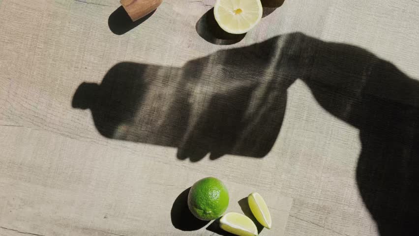 An arrangement of lemons and limes with a grater and muddler on a wooden surface. Sunlight casts a shadow of a bottle, creating an artistic composition ideal for a culinary or beverage preparation the