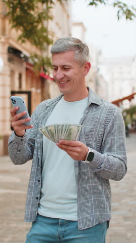 Happy rich man counting holding money dollar cash, using smartphone calculator app, plans to order gifts and food delivery online, booking hotel room. Guy tourist in city street, outdoors. Vertical.