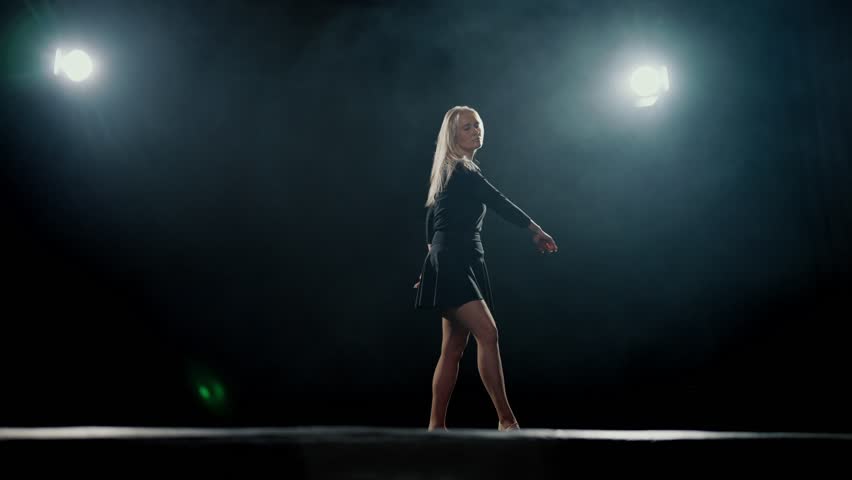 Graceful blonde woman in black dress dancing alone in dark studio, slow motion. Beautiful lady performing contemporary choreography on stage, rehearsing in dance hall, bright soffit and artistic smoke