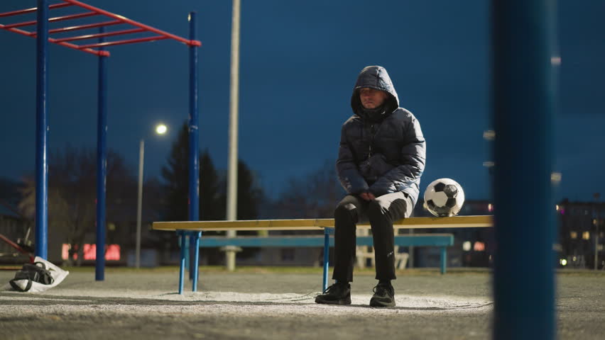 A man sits alone outdoors at night in an empty stadium, dressed in a hooded jacket with his hands clasped between his legs, with blurred views of a building and goalpost in the background