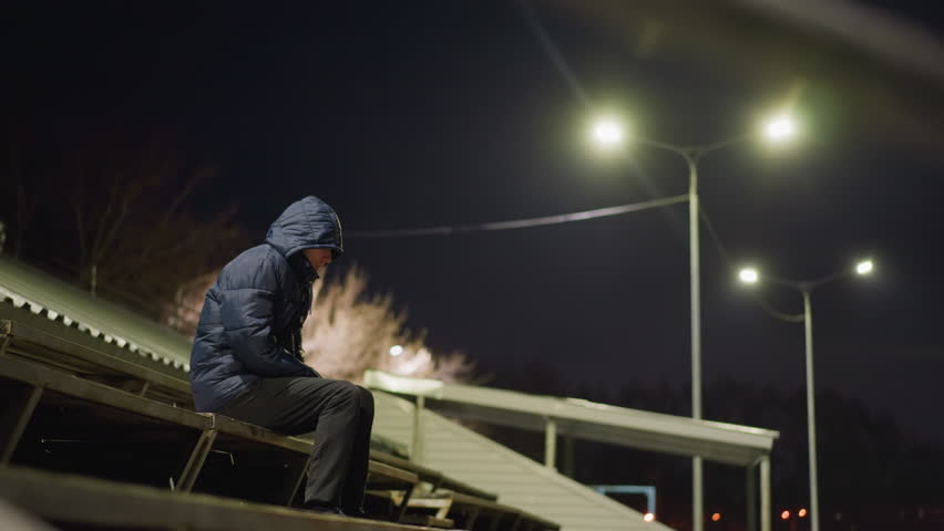 A man wearing a hood is seated alone on bleachers at night, with streetlights illuminating the surrounding area