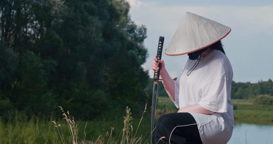 A woman with a katana in a straw hat, bending one knee, hiding her face behind a straw hat, stands on the edge of a cliff by the river. Beautiful cosplay of a samurai with a katana and a straw hat.