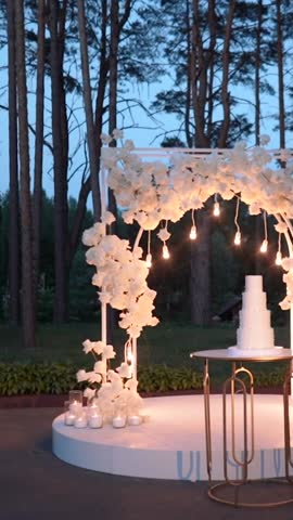 Evening wedding arch of white flowers and greenery with lanterns outdoors. Floristry and decor at the solemn ceremony. Large white cake with cream