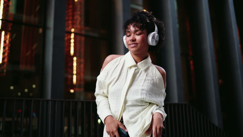 African American Woman dancing outdoors while listening to playlist on her smartphone and wireless headphones. Carefree female enjoying freedom of movement and music streaming through mobile app