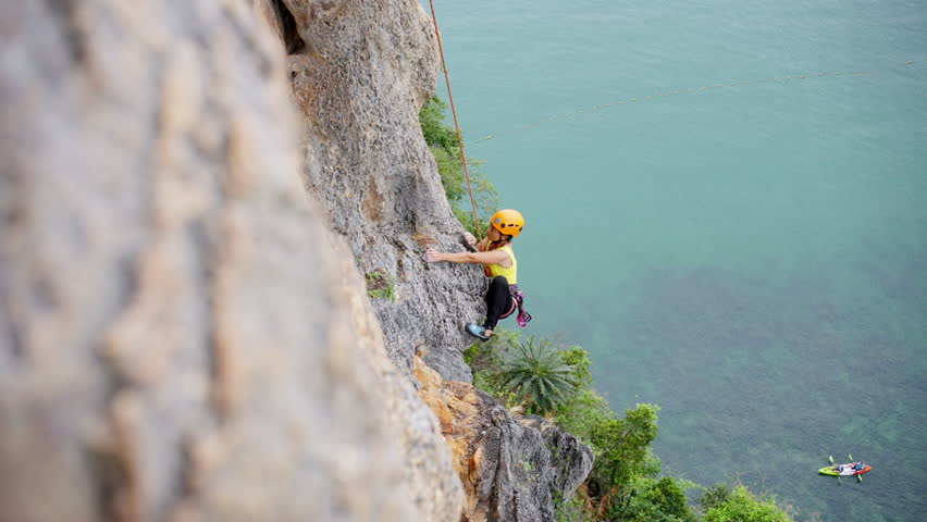 Extreme sport climbing on summer beach holiday vacation. Healthy Asian mature woman enjoy outdoor active lifestyle climbing on rocky mountain with safety equipment and hanging rope at tropical island.