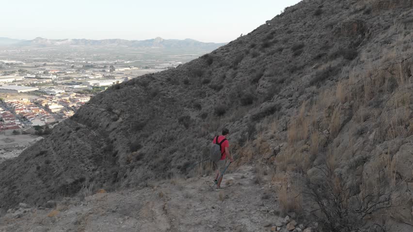 A man with trekking poles, in shorts and a red shirt, climbs steep rocks off-trail, seeking the summit and success in the morning sunlight.