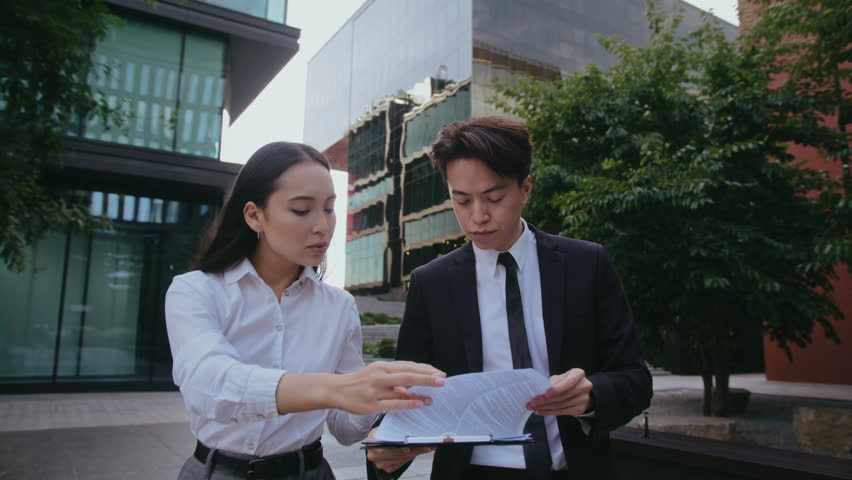 Asian business partners man and woman discuss plans examining documents on clipboard in business district. Businesspeople prepare presentation data walking about downtown