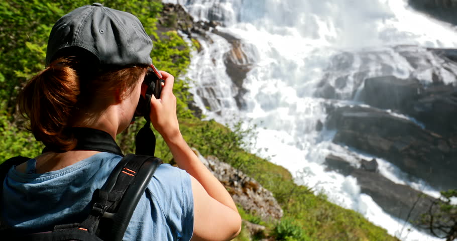 Kinsarvik, Hordaland, Norway. Young Woman Lady Tourist Traveler Taking Pictures Of Waterfall Nyastolfossen In Hardangervidda Mountain Plateau. Nyastolsfossen Is Famous Popular Destination.