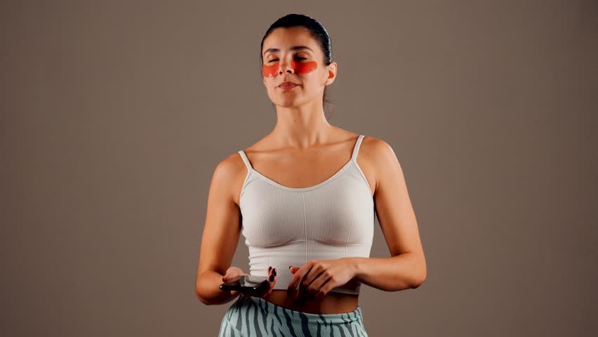 Young woman with perfect glowing skin and natural makeup posing for the camera with medical collagen patches under her eyes to reduce fatigue and puffiness. Skin care.Modern beauty tools concept 4k