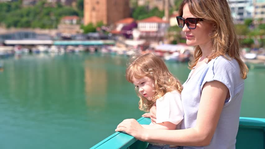 Young woman in sunglasses and curly blonde baby, leaving sea bay on cruise ship, standing on deck