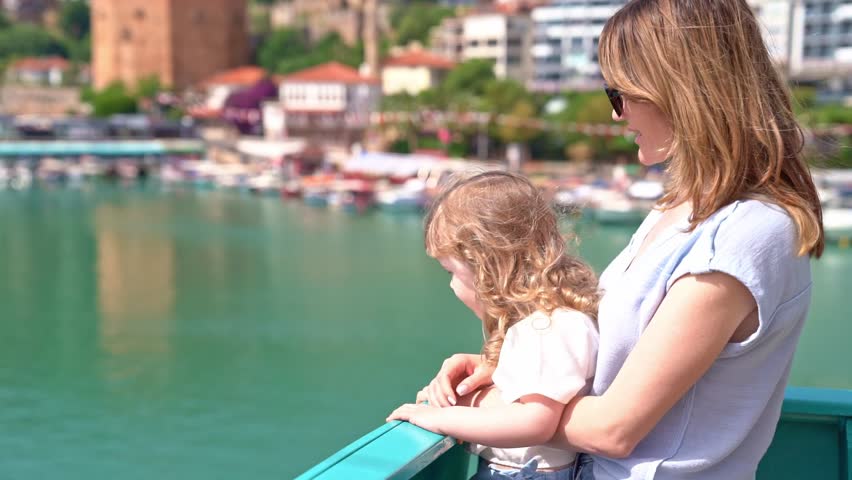 Young mother and little curly blonde daughter on deck of cruise ship, leaving sea bay on sunny summer day, admiring panorama, family trip conceptl.