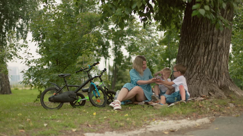 A mother sits with her two children under a tree in the park, offering something to her older son, who appears to refuse the item, the younger child seems more interested
