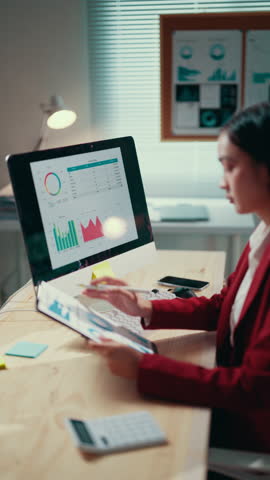Businesswoman in her office is reviewing data displayed on a computer screen and a tablet, using a pen to point at specific information on both devices