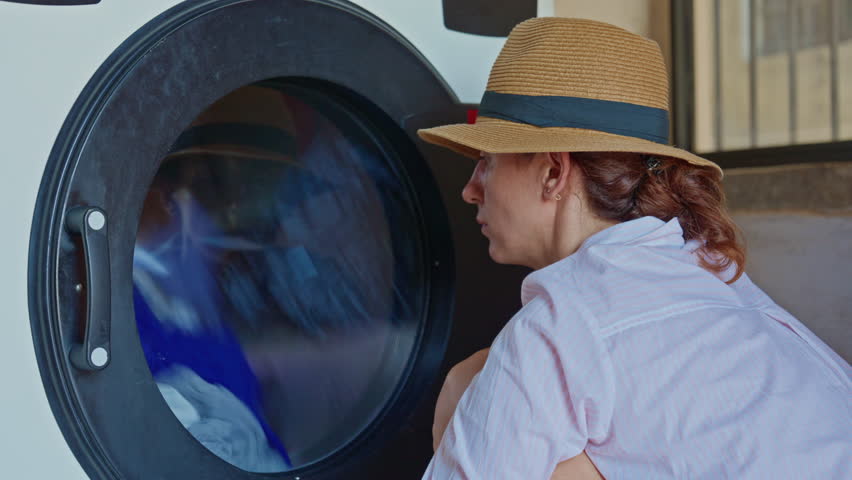 Woman tourist in hat sitting in laundry room waiting for washing to finish