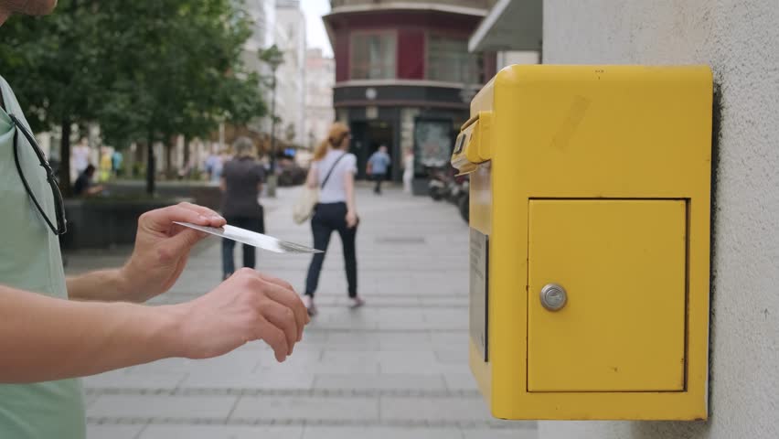 Male hand drops postcard in a yellow postbox. Man tourist sending post card and put it in yellow street mailbox. Postcrossing hobby concept
