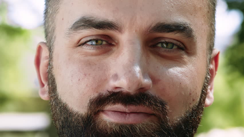 A man with a beard and striking green eyes smiles warmly in a natural outdoor setting.