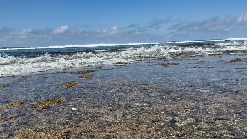 slow-motion waves crashing onto a rocky shoreline. The water splashes and foams as it reaches the shore, with the clear sky and distant horizon adding to the peaceful and natural scene