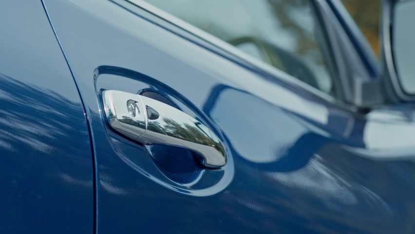 A hand reaches out to open the door of a blue car while parked outdoors under bright sunlight.