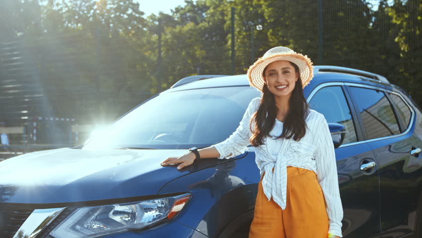 A young woman smiles while leaning against a blue SUV in a bright and sunny environment.