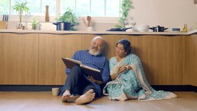 Joyful Indian Asian senior couple sitting comfortably on the floor of their modern, well-equipped kitchen relaxing with cups of tea, laughing together as they look through an old photo album - Powered by Shutterstock - Get 15% off with code: PIKWIZARD15