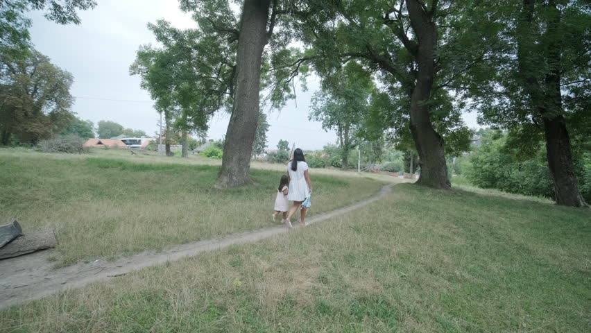A pregnant woman and her young daughter, both dressed in white, walk hand-in-hand through a grassy park. The scene captures a serene moment of family bonding in nature, surrounded by trees.