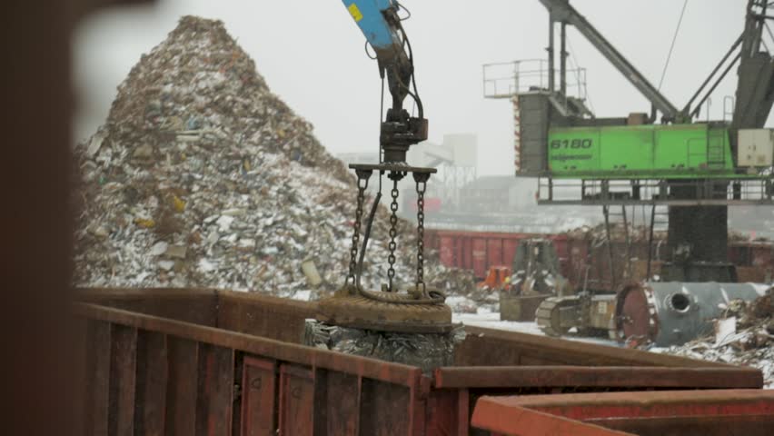 A magnet crane lifts crushed metal scrap in an industrial recycling yard on a snowy day