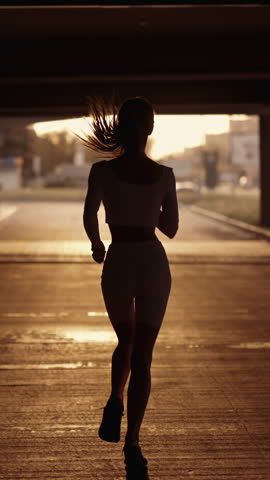 A young sporty woman runner is listening to music in earphones and training. Slim fitness girl is jogging outdoor. Concept of workouts running and healthy sport lifestyle. Vertical video footage