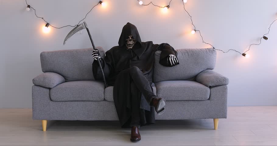 Funny man in Halloween grim reaper death costume holding scythe in black cloak sitting on sofa on gray background with festive garland. Halloween party and horror concept. 4k video.