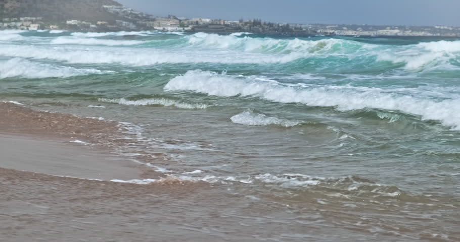 Side view, turquoise ocean waves in motion roll onto shore in morning light. Water with captive white scallops. Long waves coming shore on sunny day. Blue wave with white foam coming on Greek island