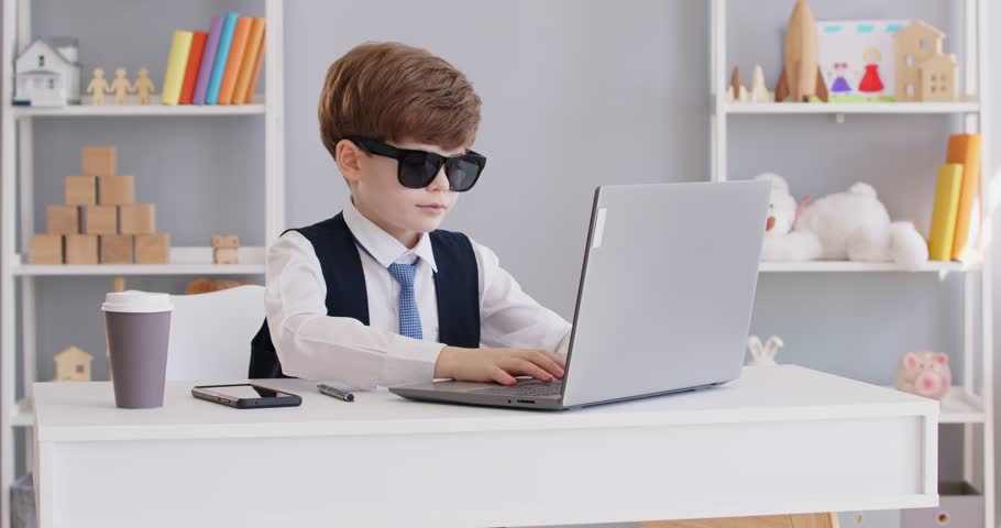 Funny preschool child boy businessman wearing suit and sunglasses working on laptop sitting at desk in office and talking by mobile phone. Little cute boss entrepreneur working on workplace. 4k video