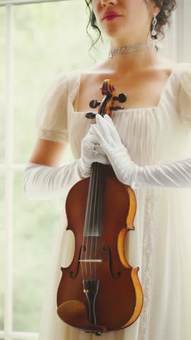 Fashion model woman musician violinist holding in hands wooden violin old style. Girl standing on sill window view on green garden. Sexy beauty face White vintage long dress. art vertical video crop.