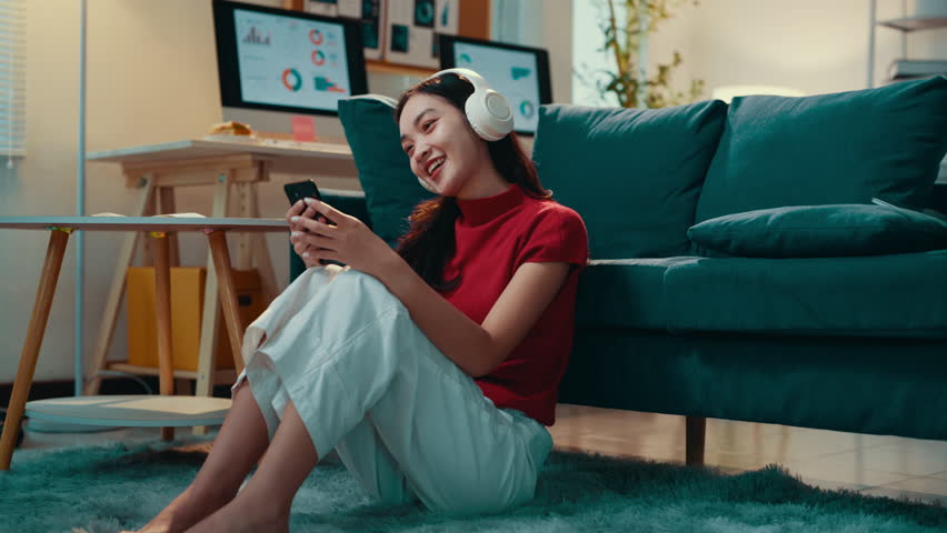 Young woman is sitting on the floor in her living room, wearing headphones and listening to music on her phone. She is smiling and enjoying herself